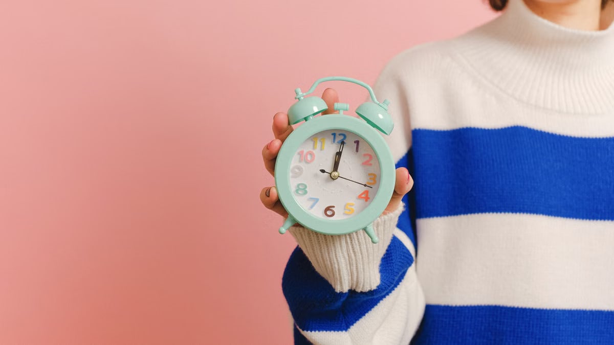 A person wearing a striped sweater holds a pastel alarm clock, against a pink backdrop.